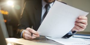 Portrait of modern bearded businessman holding papers in hands, reading and analyzing contract documentation at desk with laptop